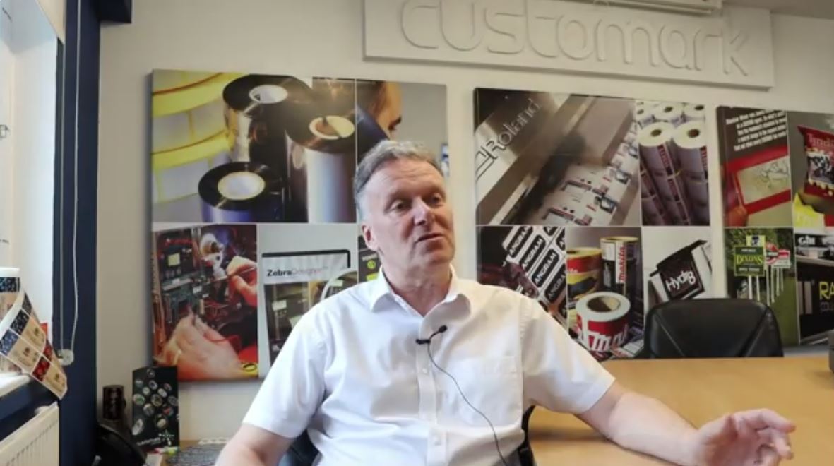 Man being interviewed with the words Customark behind him and images of print production