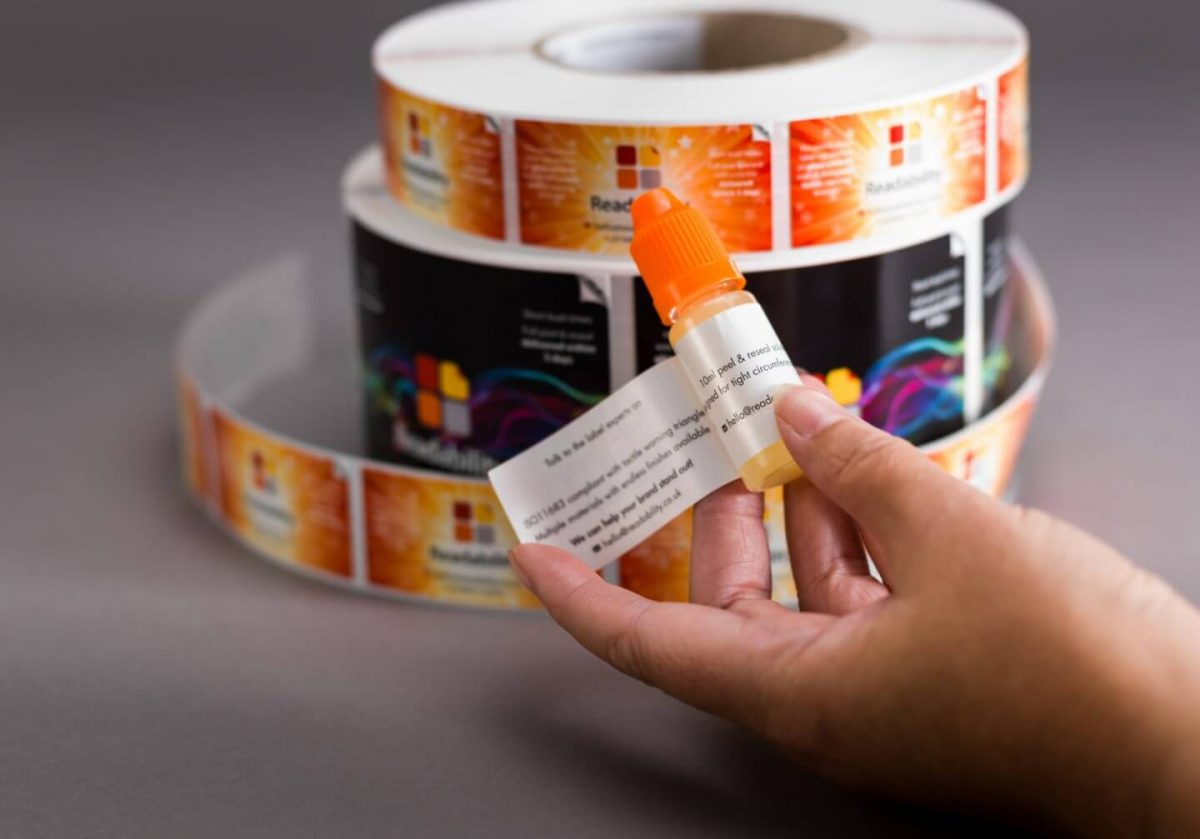 A roll of peel and reveal labels sits in the background, in the foreground a hand holds a bottle of liquid and on it the label is being demonstrated